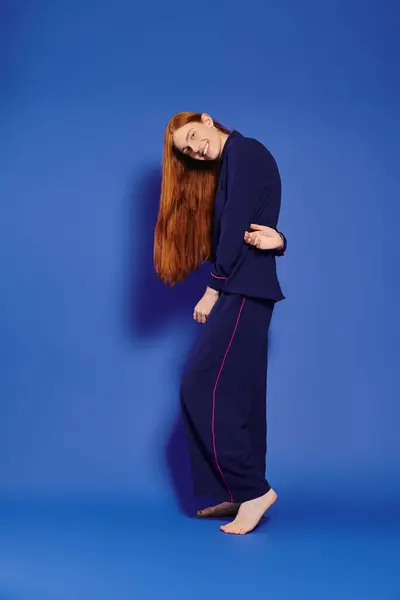 A fashionable queer man with stunning long red hair stands barefoot against a bold blue backdrop. — Stock Photo
