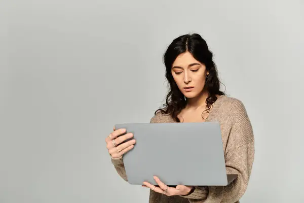 Mujer joven inspiradora se involucra con el ordenador portátil en el entorno del estudio en el fondo gris. - foto de stock