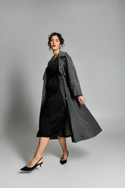 Giovane donna in abito nero e cappotto grigio posa elegantemente in uno studio. — Foto stock