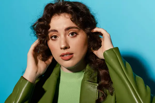 A young woman with curly hair adjusts her hair, wearing a bold green outfit in front of blue. — Stock Photo