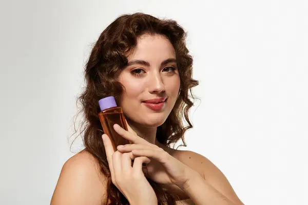Una giovane donna sicura di sé con bellissimi riccioli mette in mostra un elegante prodotto di bellezza sorridendo calorosamente. — Foto stock