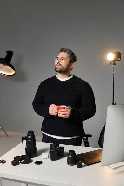 Um homem com uma camisola preta fica pensativo em uma mesa moderna cercada por equipamentos de fotografia. — Fotografia de Stock