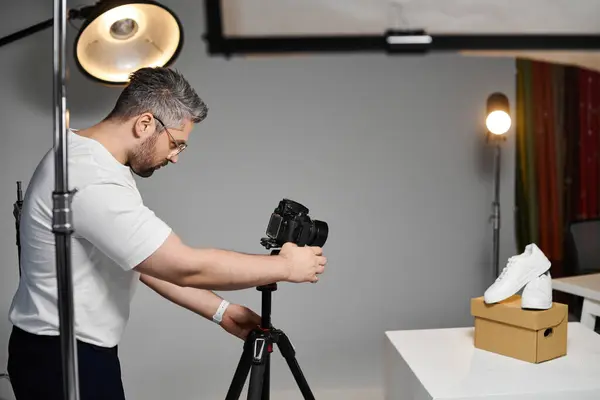 Сосредоточенный человек настраивает камеру, чтобы фотографировать стильные туфли в хорошо освещенной студии. — стоковое фото