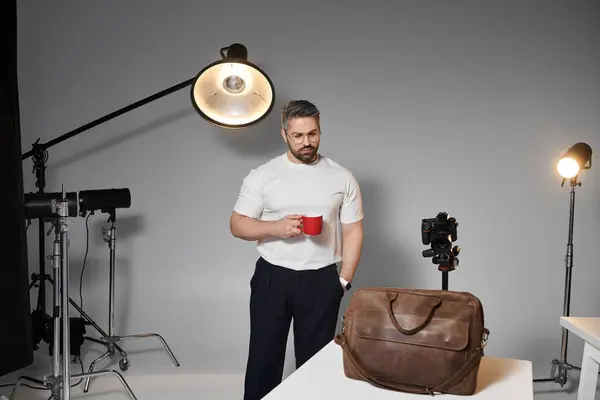 Un hombre se para pensativamente en un estudio de fotografía, sosteniendo una taza roja y rodeado de equipo. - foto de stock