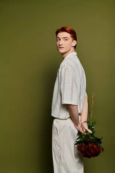 A young man hides a bouquet of roses behind his back, looking thoughtful against a green backdrop. — Stock Photo