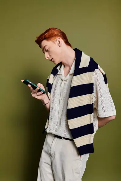 Un joven guapo se para pensativamente, mirando su teléfono inteligente, vistiendo un atuendo de moda. - foto de stock