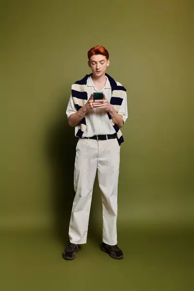 A handsome young man stands thoughtfully, focused on his mobile device, dressed in a trendy outfit. — Stock Photo