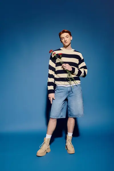 A handsome young man stands confidently, holding roses, wearing a striped sweater and denim shorts. — Stock Photo