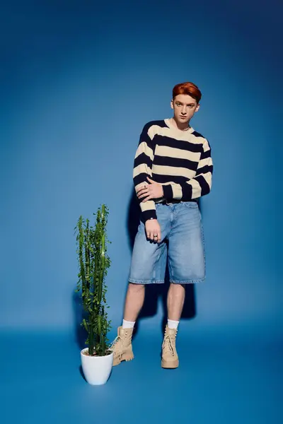 A young and handsome man stands confidently near a tall green plant, showcasing a stylish outfit. — Stock Photo