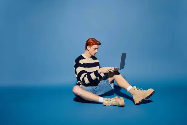 Casual young man focused on his laptop, emotionally engaged against a blue backdrop. — Stock Photo
