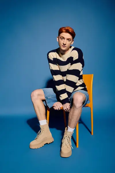 A handsome young man sits on an orange chair, showing deep emotions against a blue backdrop. — Stock Photo