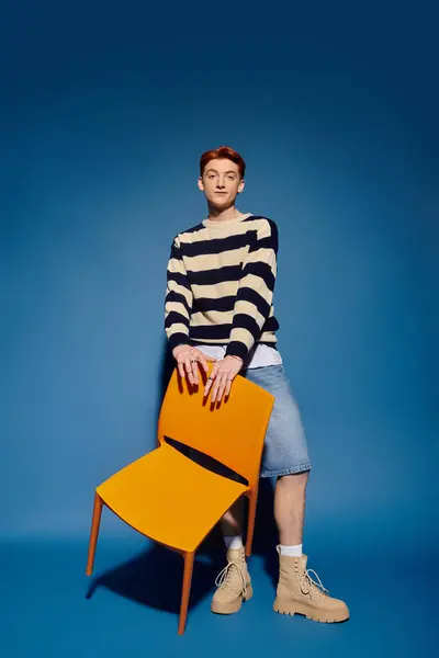A young man with striking features stands confidently beside a bright orange chair, evoking emotion. — Stock Photo