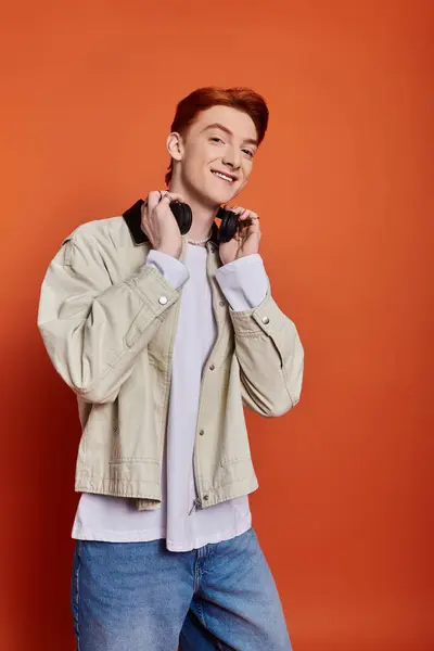 A young and charming man expresses joy while holding headphones with an orange background. — Stock Photo