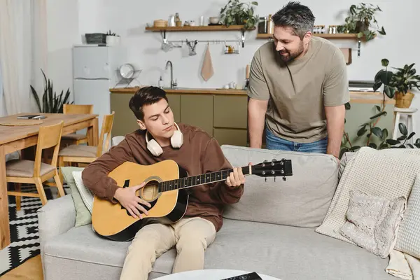 A young man plays guitar on a comfy couch while a family member watches with joy. — Stock Photo