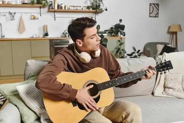 A young man strums a guitar while relaxing on a comfortable couch filled with soft cushions. — Stock Photo