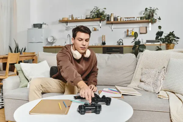 A young man relaxes on a plush sofa, gaming with controllers amid scattered creative notes. — Stock Photo