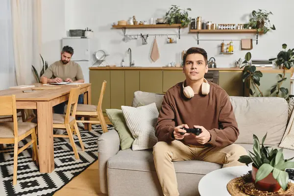 A young man enjoys gaming on the couch while another family member works on a project nearby. — Stock Photo