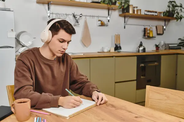 A young man sketches at a wooden table in a cozy kitchen, headphones on. — Stock Photo