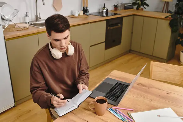 A young man sits at a wooden table in a cozy kitchen, writing in a notebook with headphones on. — Stock Photo