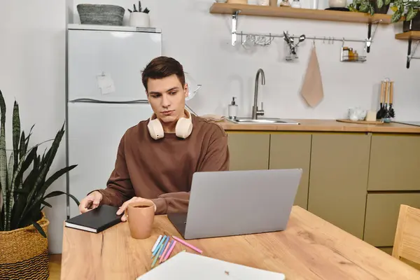 Un joven escribe en un cuaderno mientras disfruta de una bebida caliente en su acogedora mesa de cocina. - foto de stock