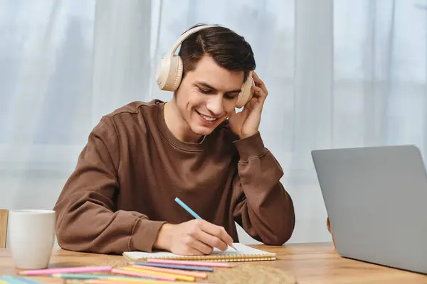 A young man is happily writing notes while enjoying music in a warm, inviting atmosphere at home. — Stock Photo