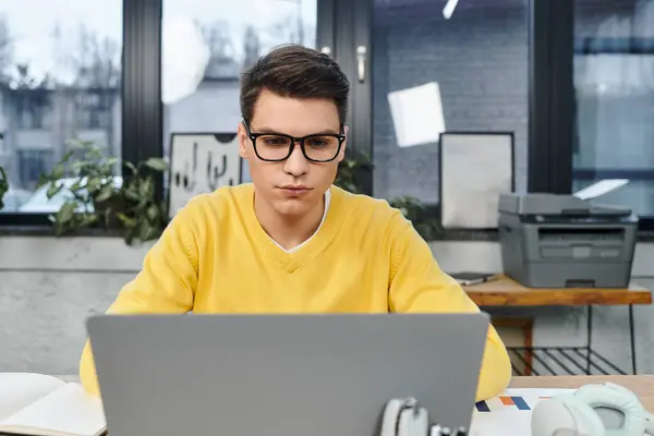 A young man in a yellow sweater concentrates on his laptop during onboarding in a bright office. — Stock Photo