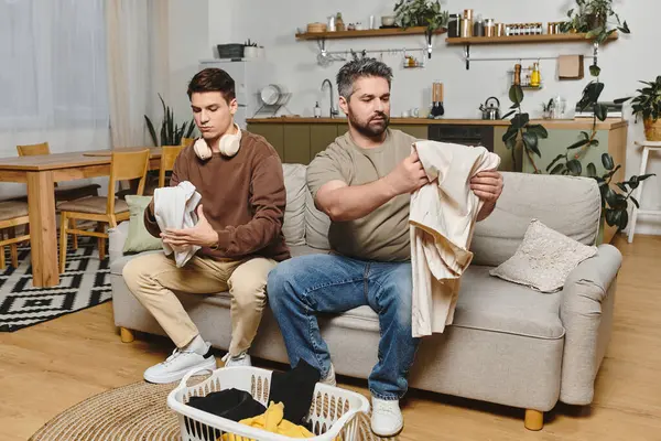 Dois irmãos dobram a roupa juntos em um sofá acolhedor, curtindo a companhia um do outro. — Fotografia de Stock