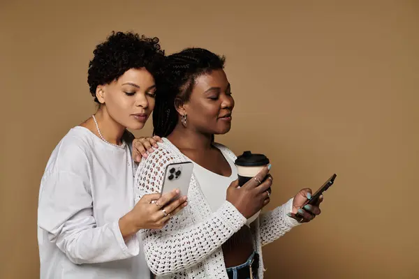 Two fashionable women share a joyful moment, engrossed in their phones and sipping coffee. — Stock Photo