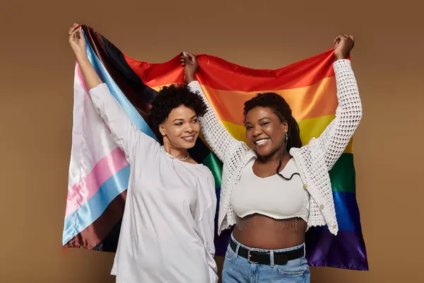 Two beautiful women proudly holding a rainbow flag, sharing joy and love in a trendy setting. — Stock Photo