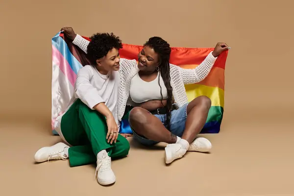 Two stylish women share smiles while wrapped in colorful rainbow flags, celebrating love. — Stock Photo