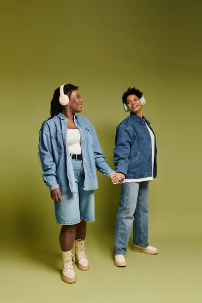 Two beautiful women in trendy denim outfits share a joyful moment while listening to music. — Stockfoto