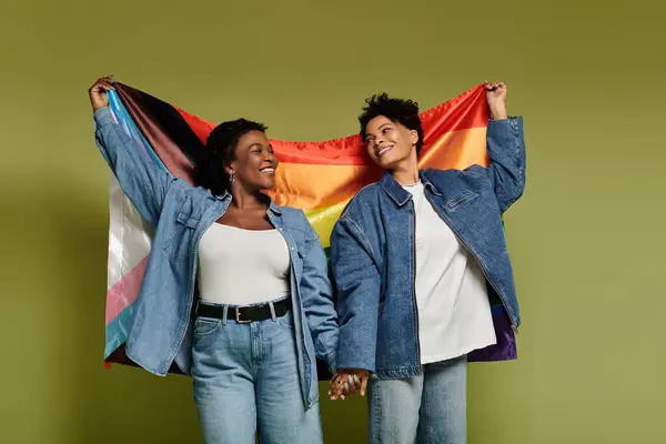 Two fashionable women hold hands and display a colorful pride flag against a bright backdrop. — Stock Photo