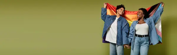 Two fashionable women hold hands and proudly display a rainbow flag against a stylish backdrop. — Stock Photo