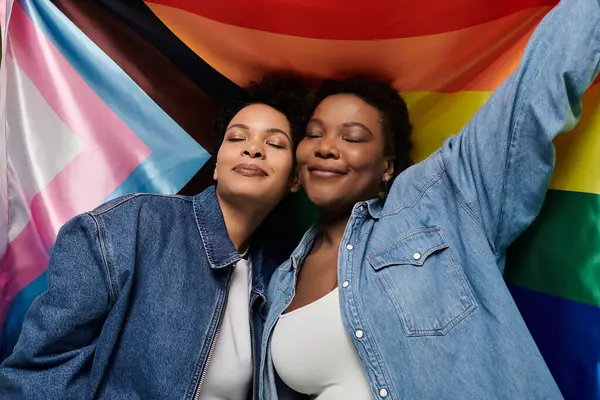 Two stylish women joyfully embrace, dressed chicly and surrounded by vibrant pride flags. — Fotografia de Stock