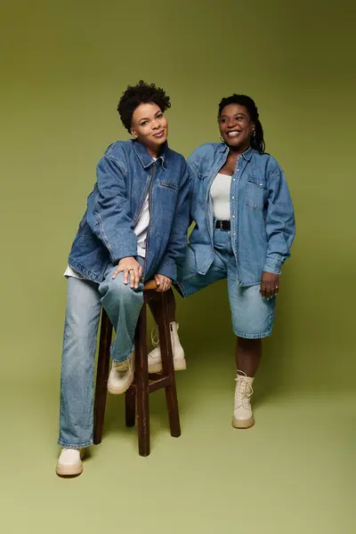 Two fashionable women pose confidently in chic denim attire against a vivid green backdrop. — Stock Photo