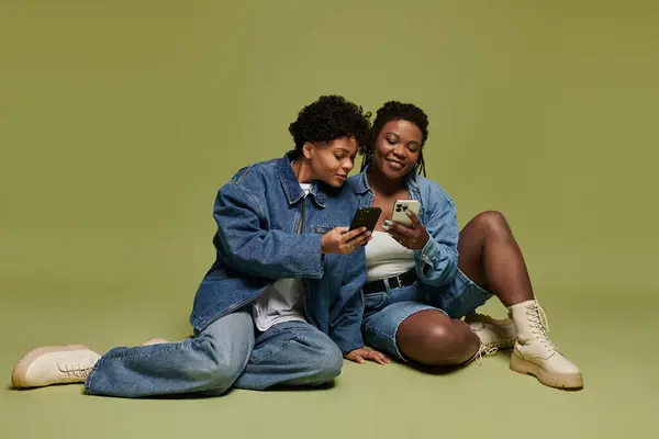 Two vibrant women share a delightful moment, casually exploring their phones in fashionable attire. — Stock Photo