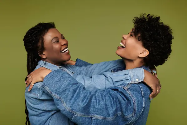 Two women in stylish denim jackets joyfully embrace and laugh against a vivid green backdrop. — Stock Photo