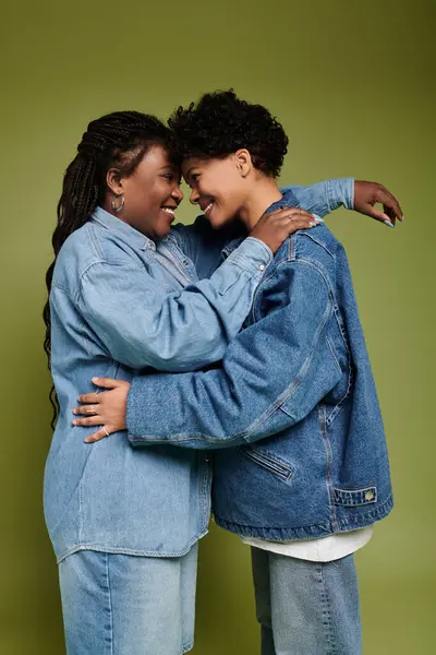 Two beautiful women in trendy denim jackets share a joyful embrace against a vivid green backdrop. — Stock Photo