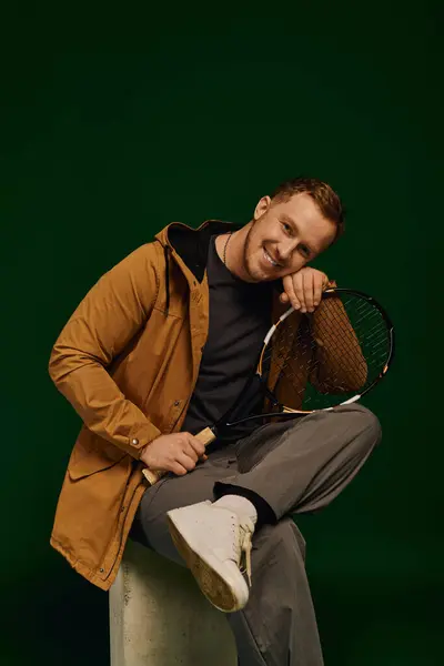 A good looking man in casual attire poses confidently with a tennis racket in a studio. — Stock Photo