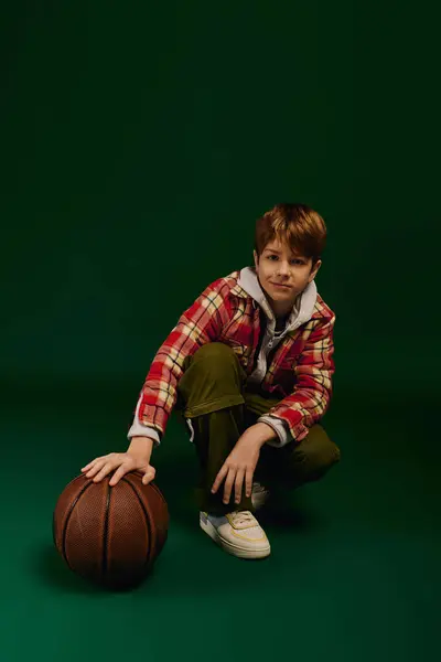 A preteen boy confidently shows off his checkered outfit next to a basketball on a green backdrop. — Stock Photo