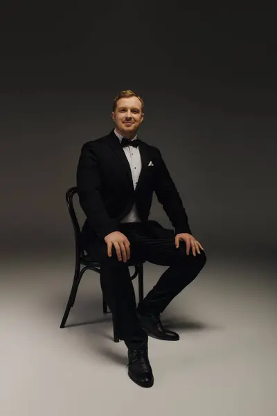 A confident man in a tuxedo poses elegantly on a chair in a sleek studio setting. — Stock Photo
