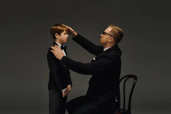 A father adjusts his son's bow tie, both dressed elegantly in tuxedos while posing in the studio. — Stock Photo