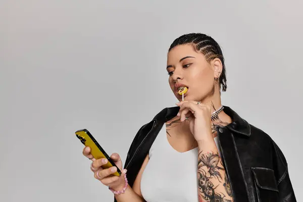 Selbstbewusste junge Frau mit Tätowierungen interagiert mit ihrem Handy und genießt einen Lutscher in einer schicken Umgebung. — Stockfoto