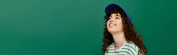 Emotional young woman wearing a cap beams with happiness while standing by a green wall. — Stock Photo