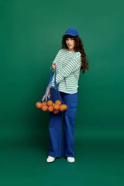 Jeune femme aux cheveux bouclés modèle une tenue tendance tout en tenant un sac d'oranges, exsudant l'émotion. — Photo de stock