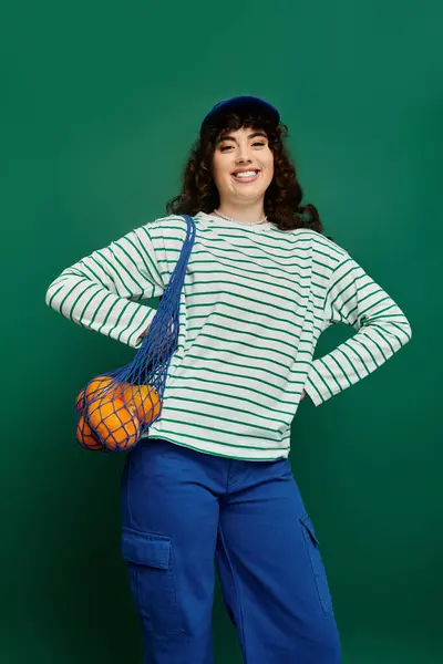 Mujer joven sonriente con una camisa a rayas y pantalones azules muestra su estilo de moda con naranjas. - foto de stock