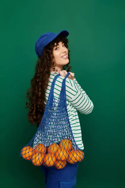 Happy woman with curly hair holds a net bag of oranges against a green background. — Stock Photo