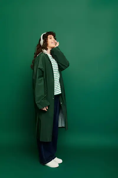 Charming young woman smiles delightfully in her trendy outfit featuring a green coat and headphones. — Stock Photo