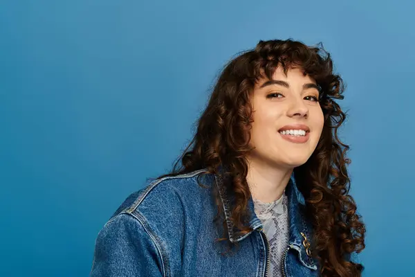 Young woman with curly hair wears a denim jacket and radiates happiness against a blue backdrop. — Stock Photo