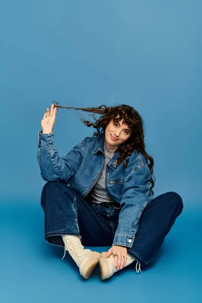 Jovem encantadora com cabelo encaracolado toca brincando suas fechaduras enquanto sentada em uma superfície azul. — Fotografia de Stock